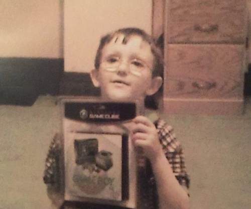 A picture of me as a 6 year old who's mildly amused, with frayed hair that goes halfway down my forhead, sporting a plaid shirt and glasses with circular lenses. Holding up an item in a plastic package with text that indicates that the item is a GameCube with a Game Boy catridge slot.